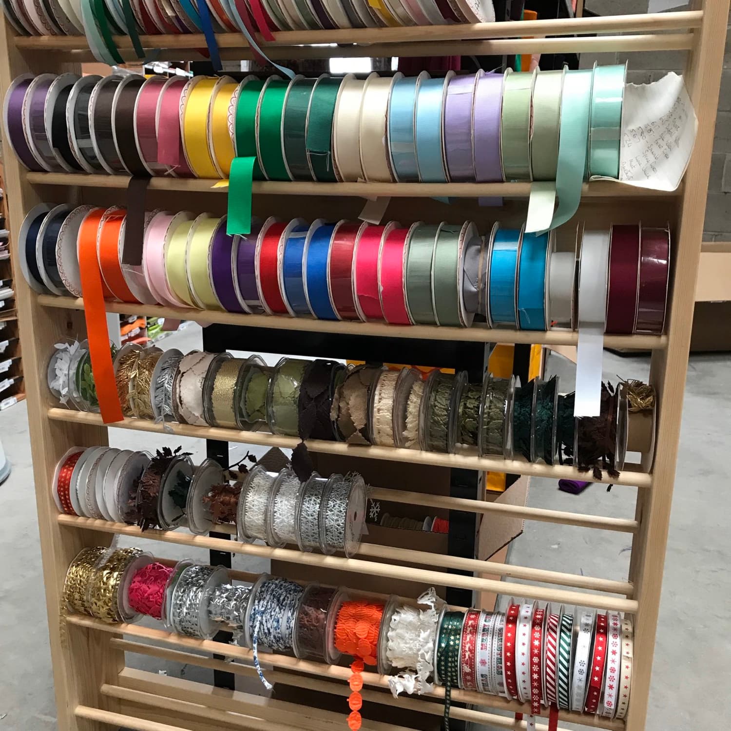 The warehouse ribbon rack, wooden dowels stacked with spools of ribbon in every colour, a handwritten stock list tucked into the top.