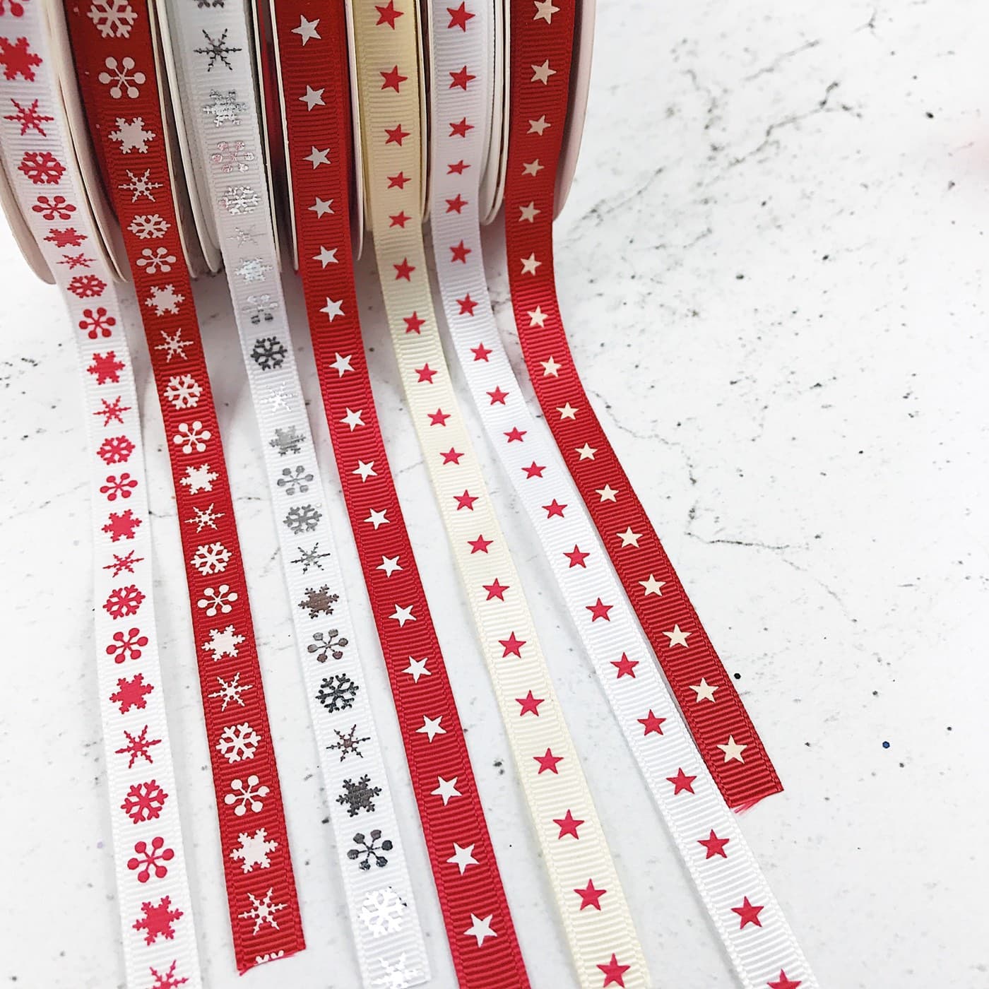 Spools of red, white and cream Christmas ribbon printed with snowflakes and stars, unspooled across a worktop.