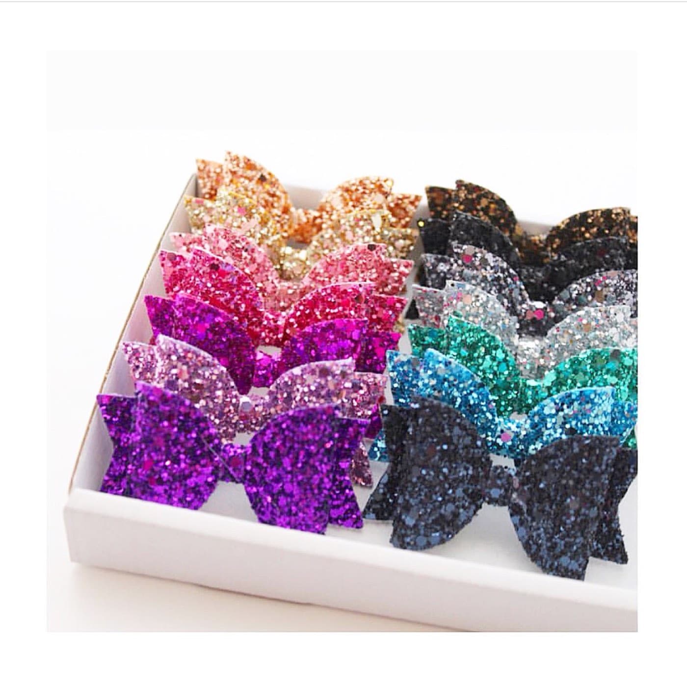 A box of handmade glitter hair bows in rows, gold, pink, purple, teal and navy.