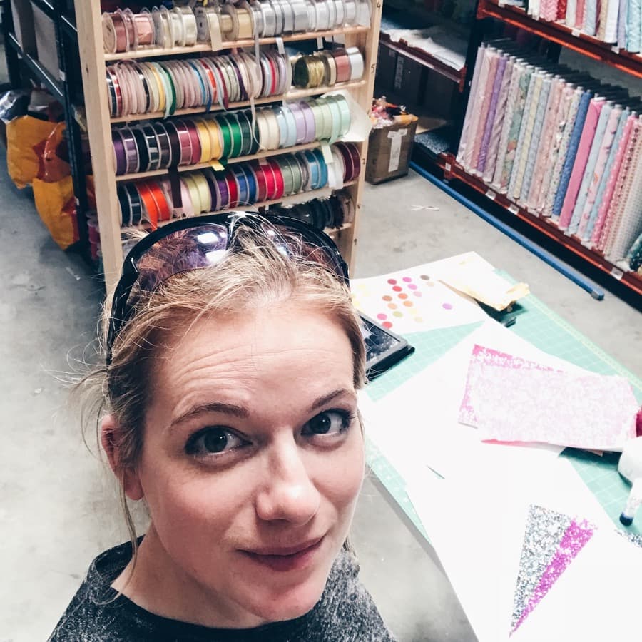 Kerri at her cutting table in the warehouse, ribbon spools racked behind her and bunting pieces cut out on the mat.