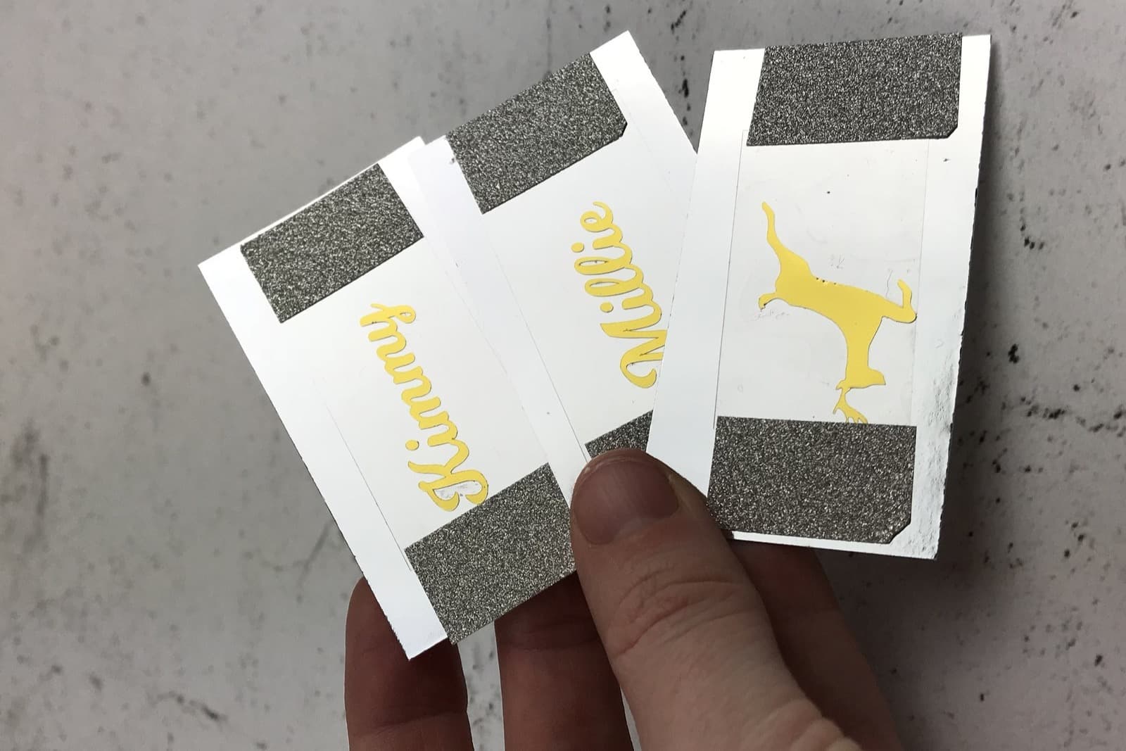 Three white vinyl name decals held in a hand against a pale concrete wall, two reading Kimmy and Millie in yellow script.
