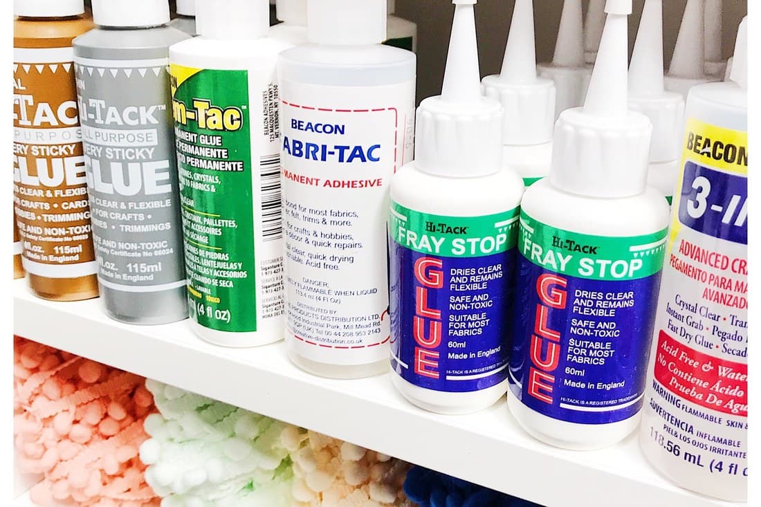 A workroom shelf of fabric glues and adhesives above trays of coloured pom pom trim