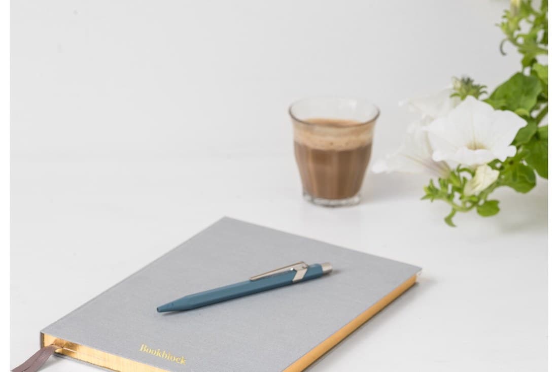 A grey notebook and pen on a white table with a glass of coffee and white flowers