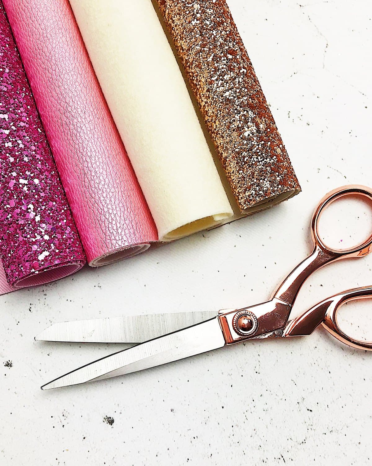 Rolls of glitter and leatherette fabric beside rose gold dressmaking shears on a white worktop.