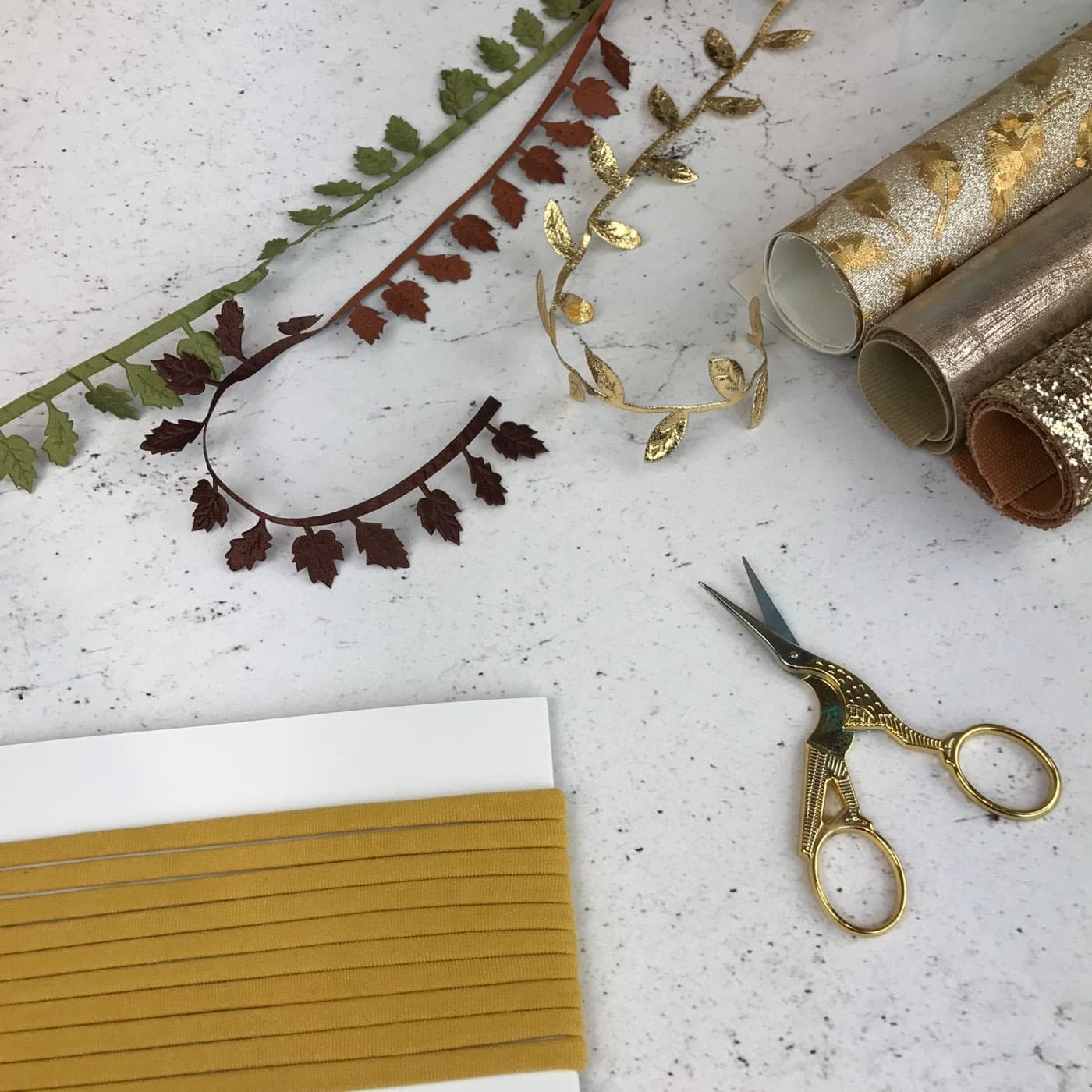 A working flatlay on marble: gold stork embroidery scissors, leaf ribbon trims and rolled metallic fabric.